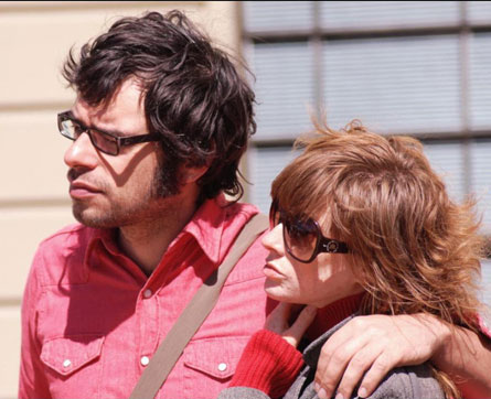 Jemaine Clement in a pink shirt hugging his wife in a grey outer.